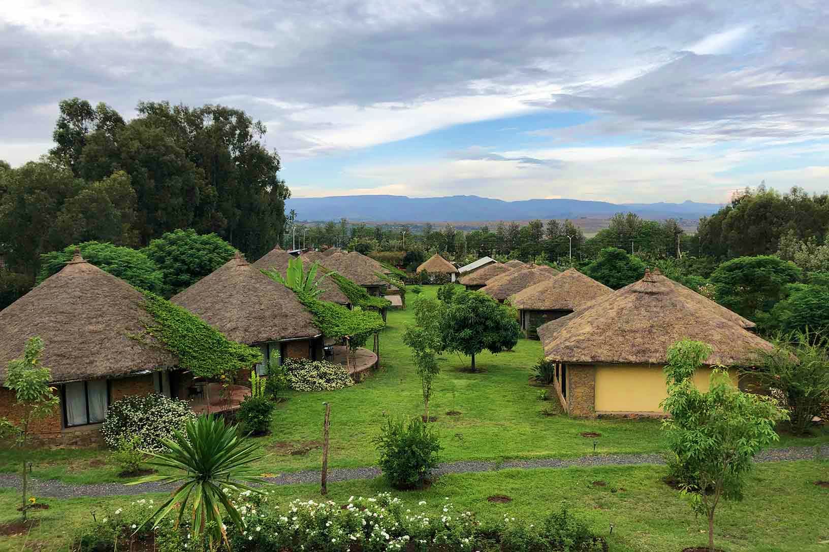 Traditional thatched-roof bungalows in lush green grounds with mountain landscape in background, Ethiopia